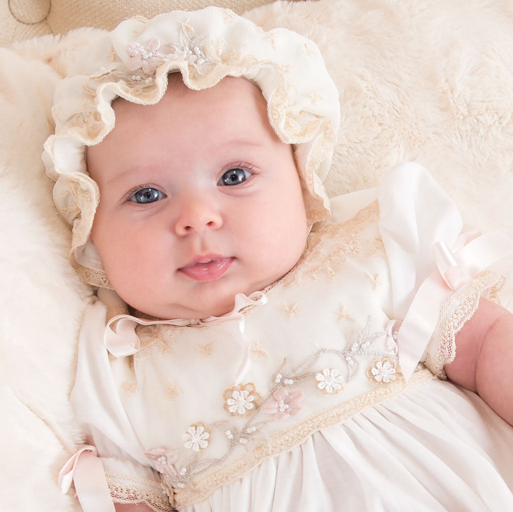 A baby with bright blue eyes lies on a soft cream blanket, wearing the Jessica Newborn Gown & Bonnet. The gown is adorned with floral appliqué and matches the frilly bonnet.