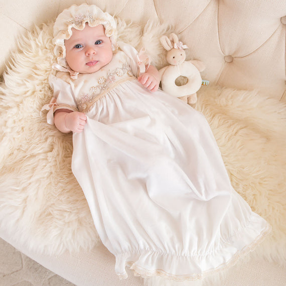 A baby wearing the Jessica Newborn Gown & Bonnet with golden embroidery and floral appliqué, lying on a fluffy white blanket, holding a soft toy rabbit, and looking up.