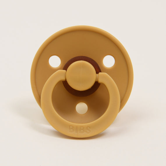 A Bibs Pacifier in Mustard yellow natural rubber baby pacifier with a circular shield and handle, along with three ventilation holes, placed against a white background.