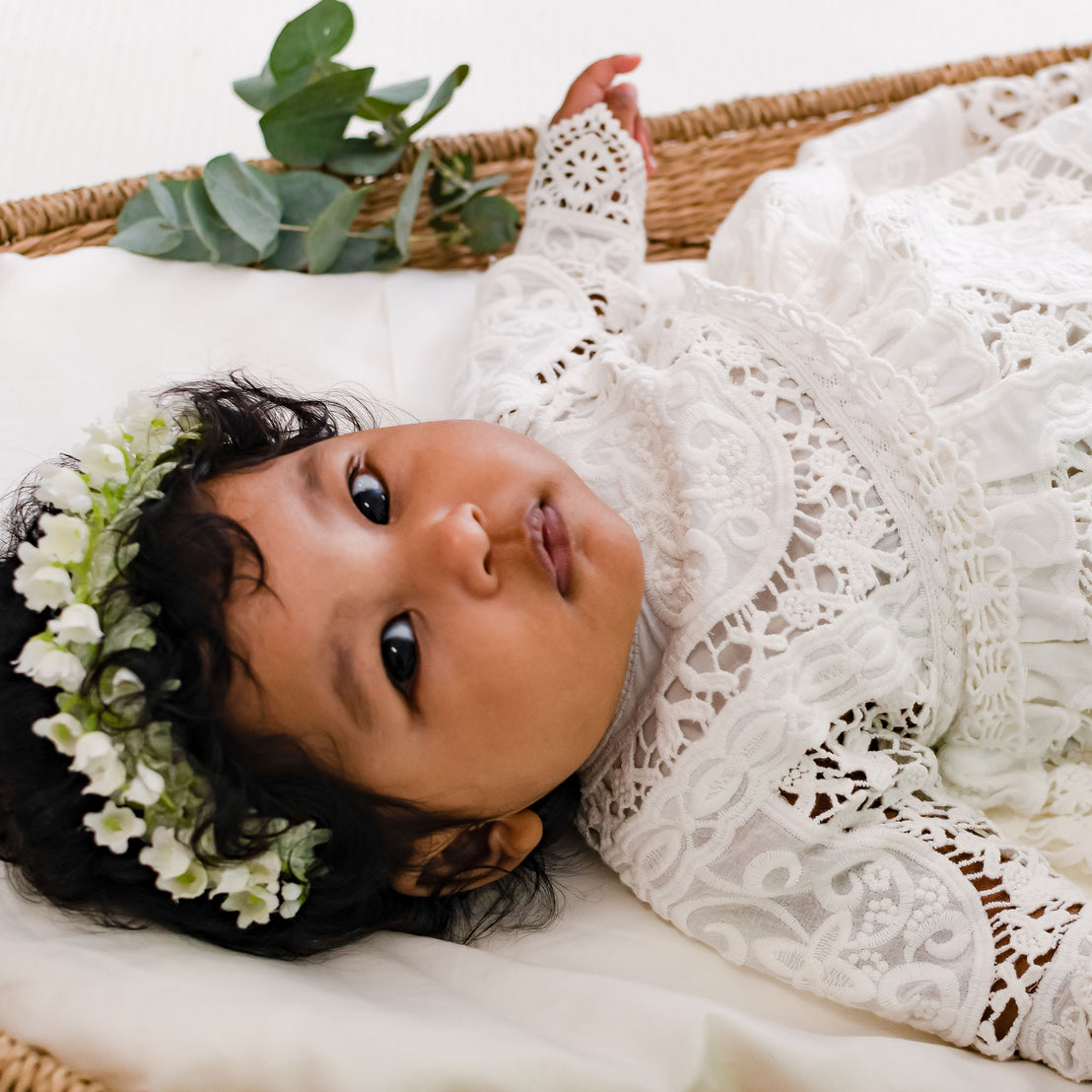 A baby with dark hair adorned in baby jewelry and wearing the Adeline Newborn Lace Dress and Bloomers complemented by a green and white floral crown lies in an upscale basket, looking up with a curious expression.