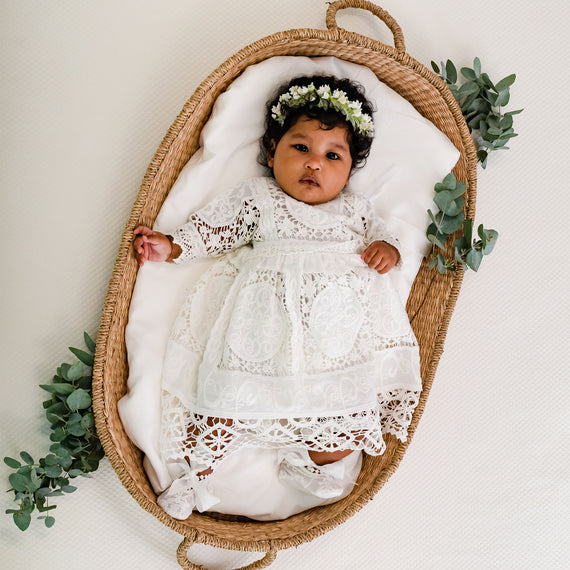 A baby wearing the Adeline Newborn Lace Dress or Gown & Bloomers lies in an upscale woven basket, surrounded by green leaves, with baby jewelry adorning it and looking upward. The baby has a floral wreath on their head.