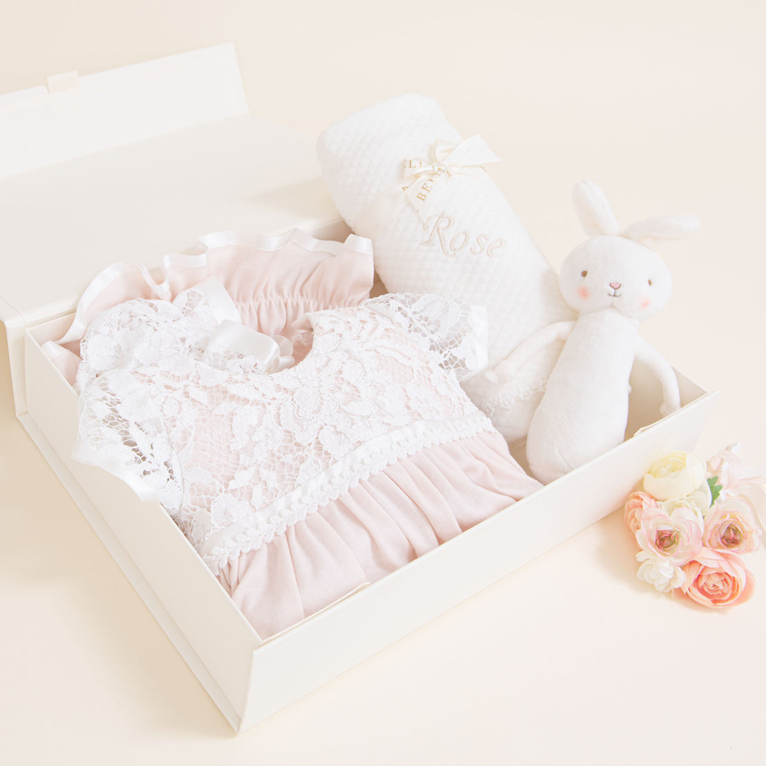 An elegant, traditional Rose Newborn Gift Set containing an heirloom lacy pink dress, a plush white bunny, and a decorative white personalized blanket with "rose" embroidered on it, displayed on a soft peach background