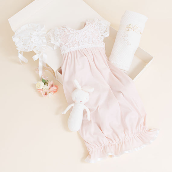 A pink heirloom baby dress with white lace details on top, displayed alongside a Rose Newborn Gift Set, a pair of baby shoes, and a small bouquet of flowers on a light cream background.