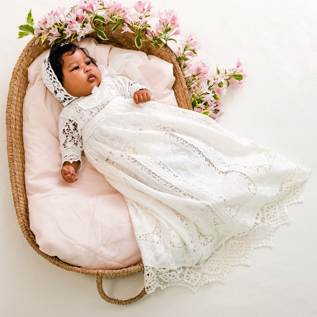 A baby in the Adeline Newborn Lace Dress or Gown & Bloomers, adorned with upscale baby jewelry, lies in a woven basket, surrounded by pink flowers, on a white background.