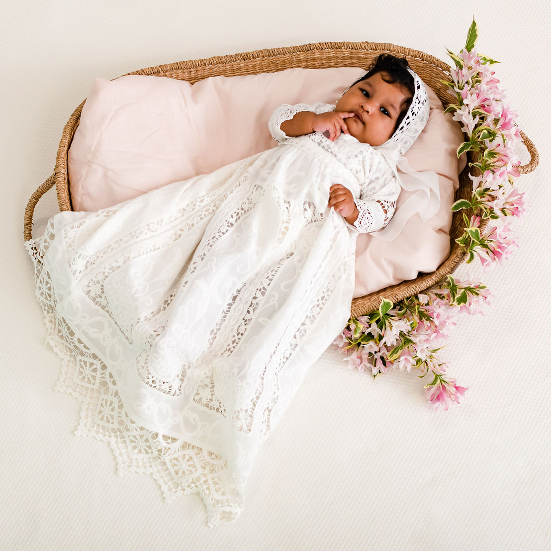 A newborn baby wearing the Adeline Newborn Lace Dress lies in an upscale wicker basket with a pink blanket, surrounded by traditional pink flowers, looking up.