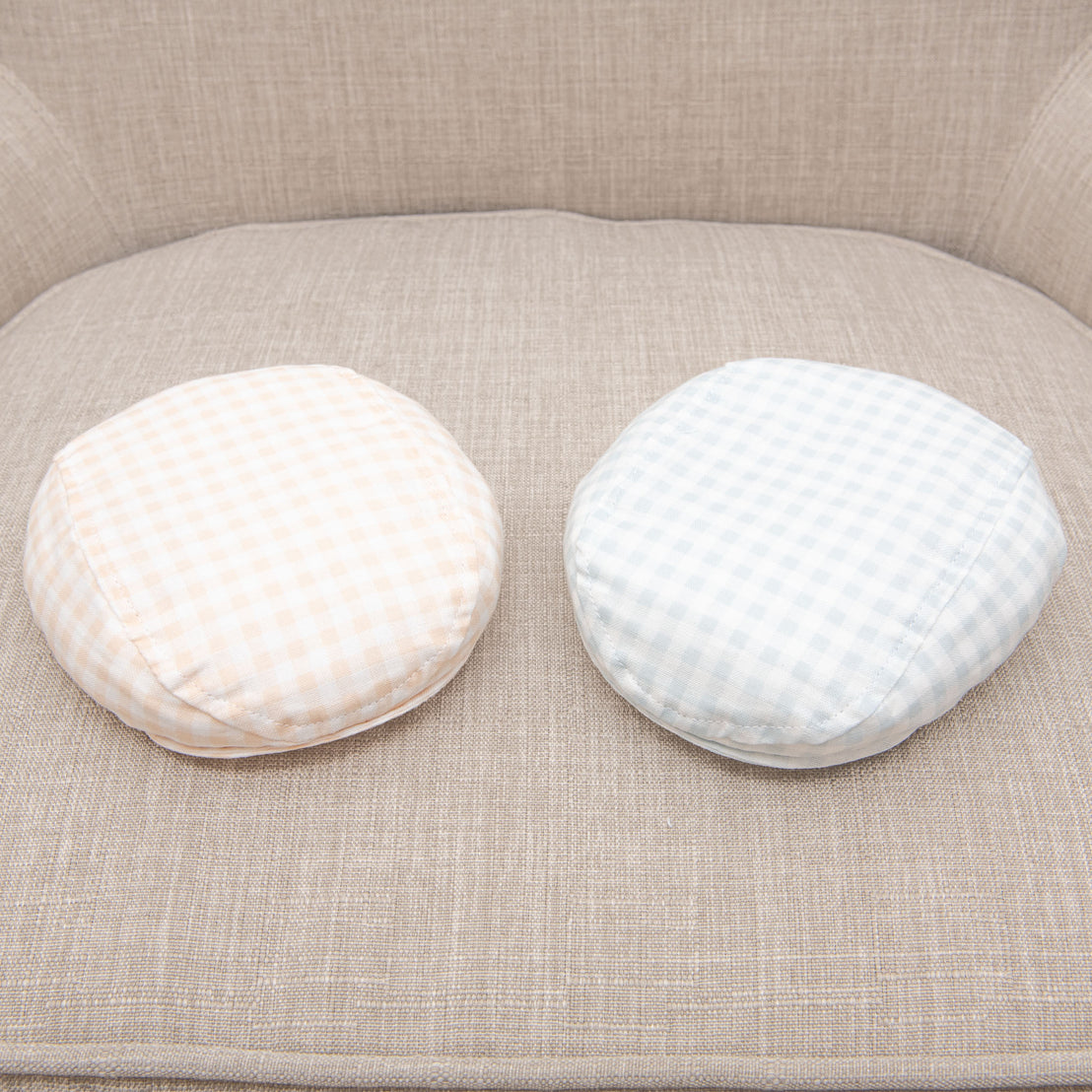 Two Ian Checked Newsboy Caps, one beige and white checked and one light blue and white checked, placed side by side on a traditional beige textured sofa.