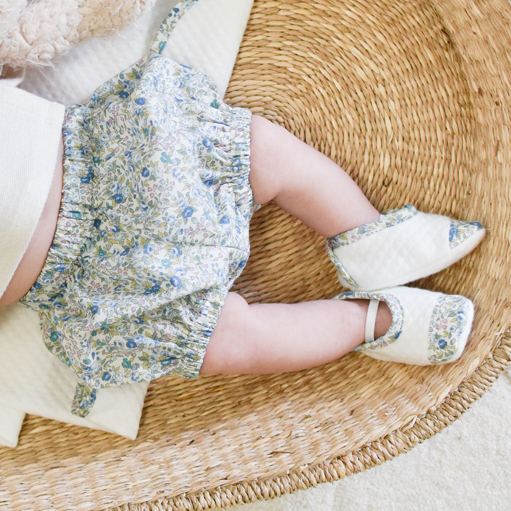 Top view of a baby’s legs wearing Petite Fleur Wrap Top & Bloomers and a floral dress, resting on a woven straw mat next to a plush bear.