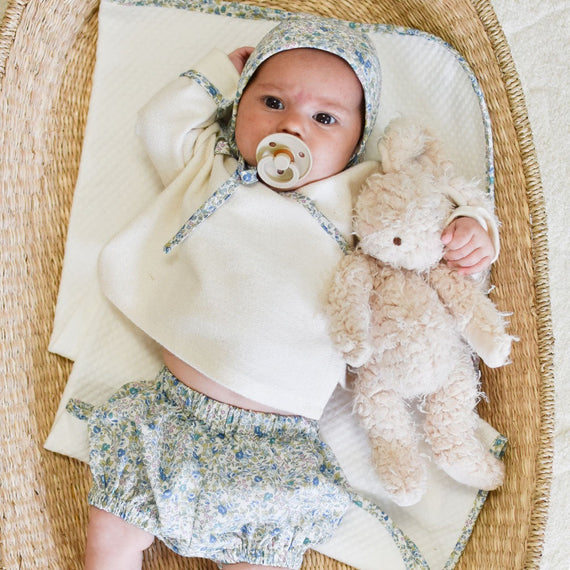 A newborn baby with a pacifier, wearing a Petite Fleur Wrap Top & Bloomers, lies comfortably in a vintage-inspired wicker basket, holding a plush teddy bear.