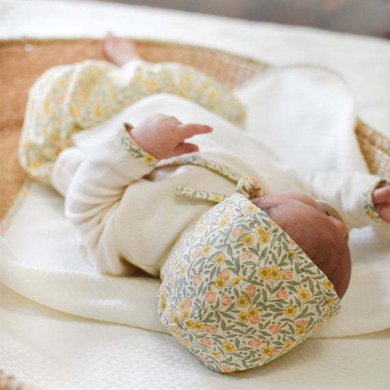 A newborn baby is lying in an upscale wicker basket, wearing a Petite Fleur Bonnet outfit with a matching hat, gently moving its tiny hands.