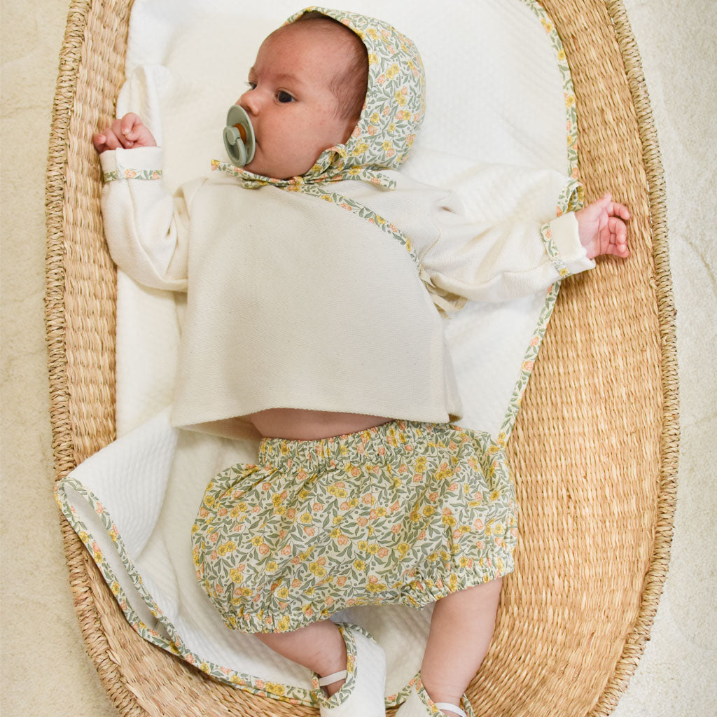 A newborn baby with a pacifier, dressed in a Petite Fleur Wrap Top & Bloomers and laying in a boutique woven basket with soft blankets.