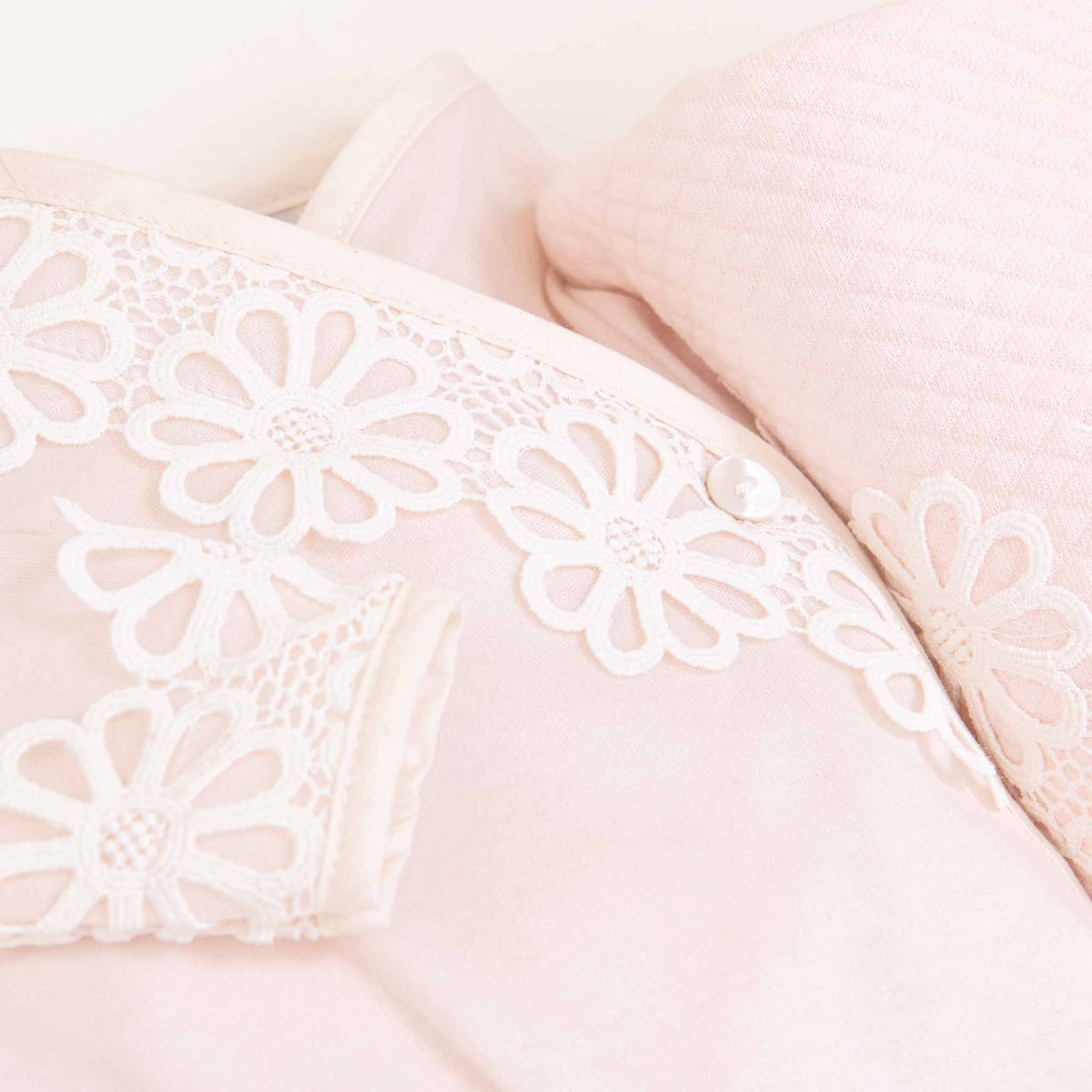 Close-up of a delicate pink Hannah Knot Gown & Bonnet from Baby Beau & Belle showcasing intricate lace details with floral patterns, against a soft, textured cushion background.