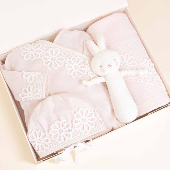 An elegant Hannah Newborn Gift Set from Baby Beau & Belle containing a white and pink soft bunny toy, a matching pink blanket, and a coming home outfit adorned with flower patterns, set against a light beige background.