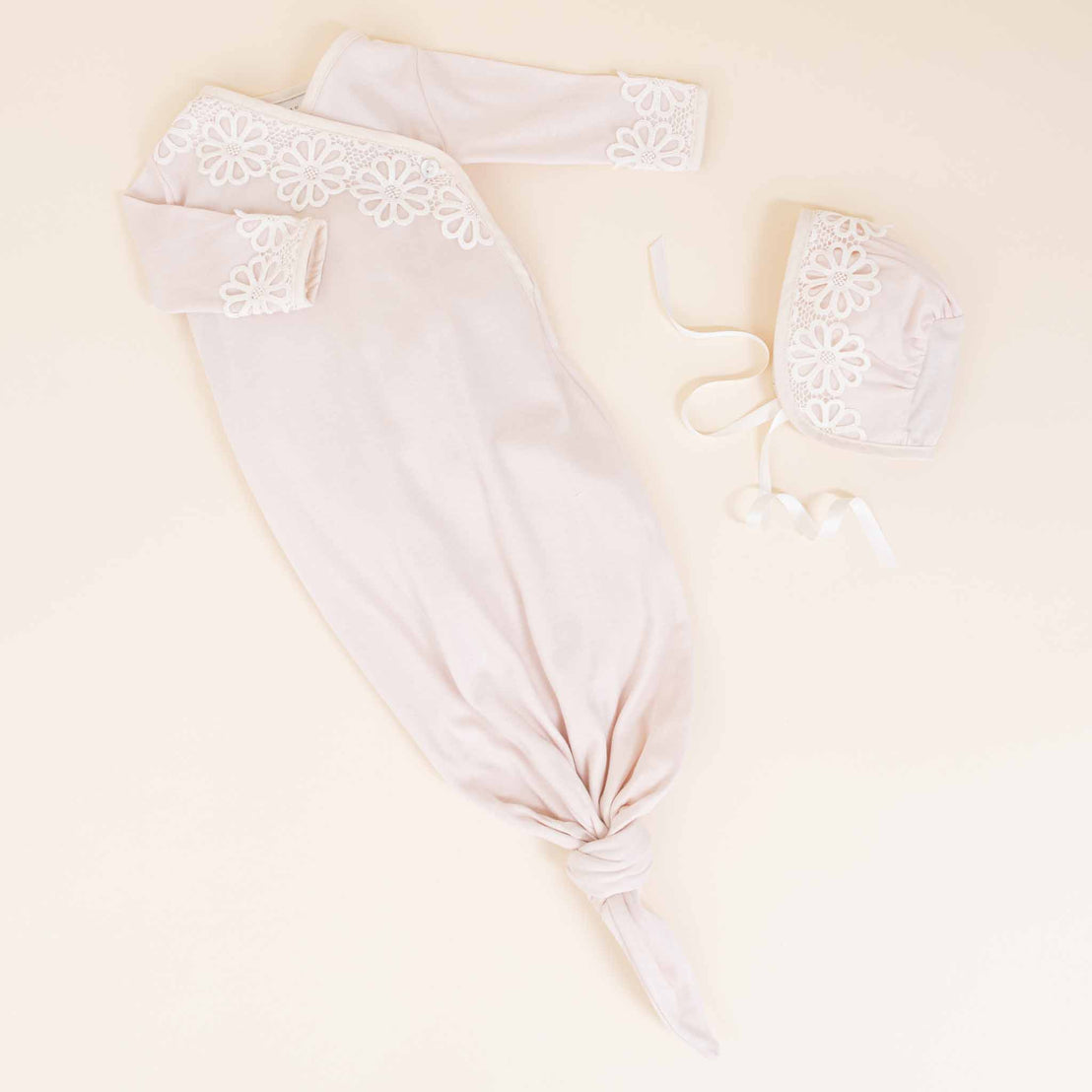 A flat lay image of the Hannah Knot Gown & Bonnet including an ivory gown with lace detailing, matching hat, and booties on a light beige background.