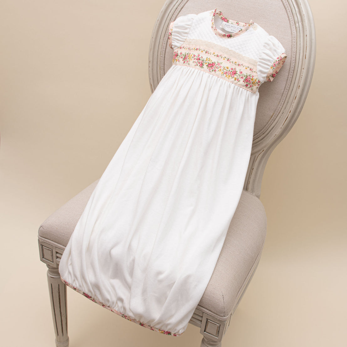 Flat lay of the Eloise Layette Gown in "Blush". Details show the white pima cotton sleeves with white quilted textured cotton bodice. The gown features cotton lace, as well as, floral and striped cotton bodice details. Button closures are on both shoulders.