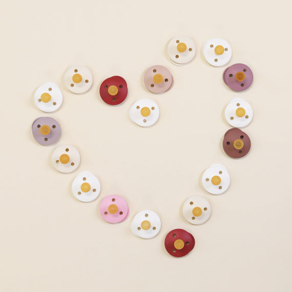 A collection of variously colored Bibs Pacifiers arranged in the shape of a heart, designed and manufactured in Denmark, on a light beige background.