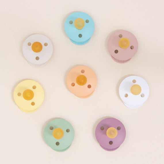 Eight colorful Bibs Pacifiers arranged in a circular pattern on a light beige background, featuring a variety of pastel shades.