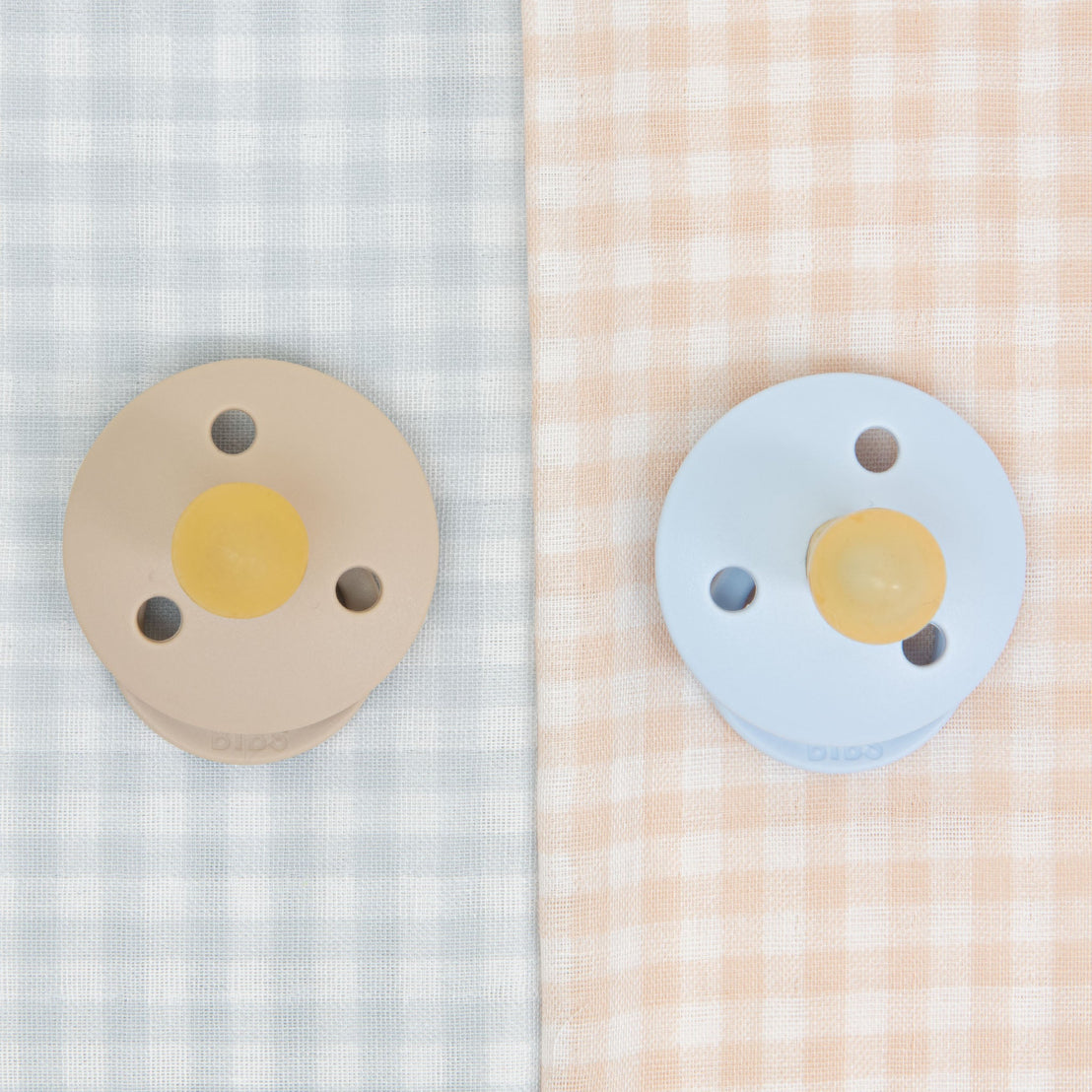 Two Isla pacifiers, one beige and one blue, placed on contrasting vintage gingham fabric backgrounds in light blue and peach colors.