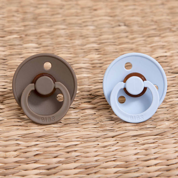 An Austin Pacifier Set in Baby Blue and Dark Oak - two baby pacifiers with a circular handle and brown button detail, perfect for a boutique coming home gift.