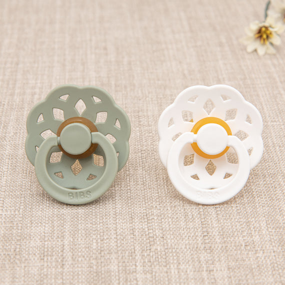 The Mila Pacifier Set on a textured fabric surface; one is sage green and the other is cream, both with a floral-like pattern around the shield.