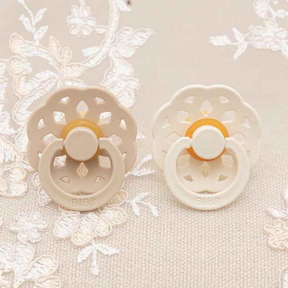 Two Kristina Pacifier Sets, one vanilla and one ivory, on a textured fabric with lace details for a christening or baptism. The pacifiers feature a unique petal-shaped design.