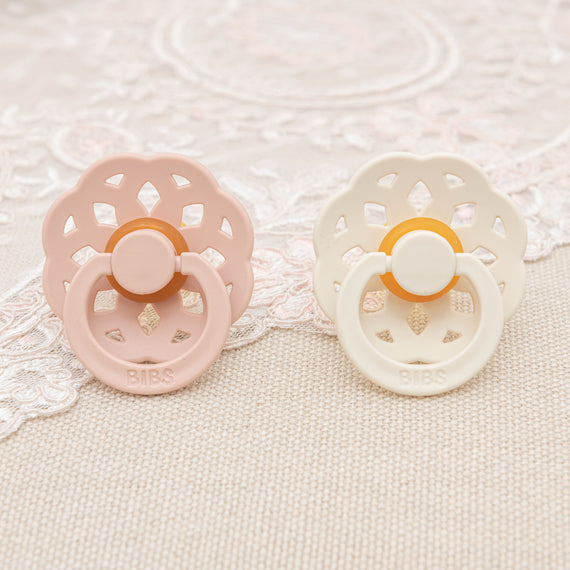 Two Elizabeth Pacifiers, one in blush pink and one in ivory, resting on a lace surface, each with a circular vintage decorative holder patterned with vent holes and a button center.