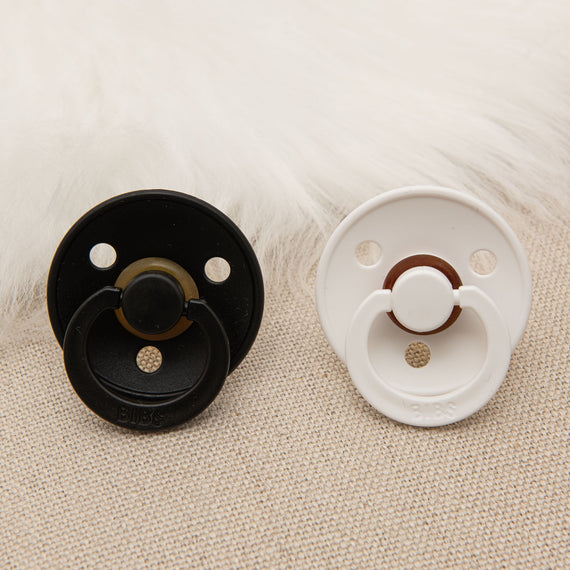 A close-up image showing a June Pacifier Set | One Black & One White baby pacifier lying on a textured fabric, with white fur partially visible in the top left corner.