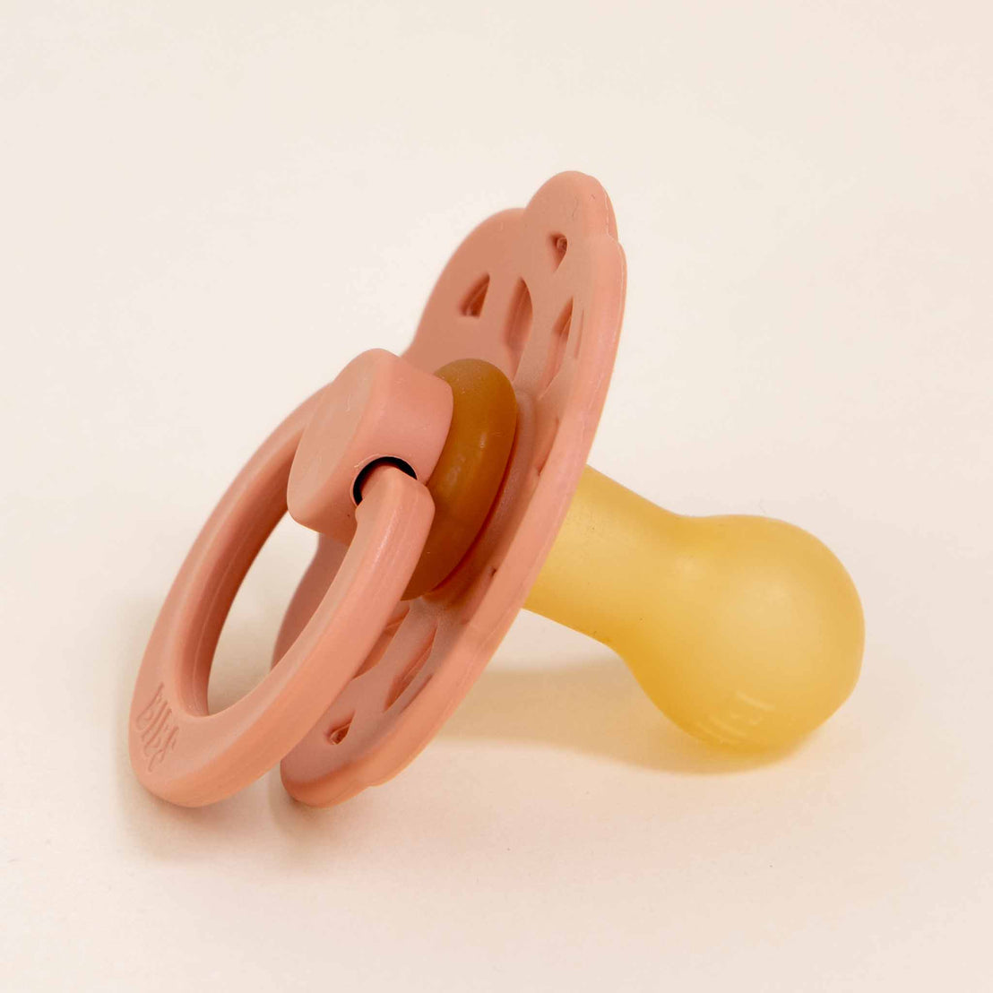 A Bibs Lace Pacifier in Peach with a handle, lying on a light beige background. The pacifier has a smooth, bulbous rubber nipple and a vented shield.