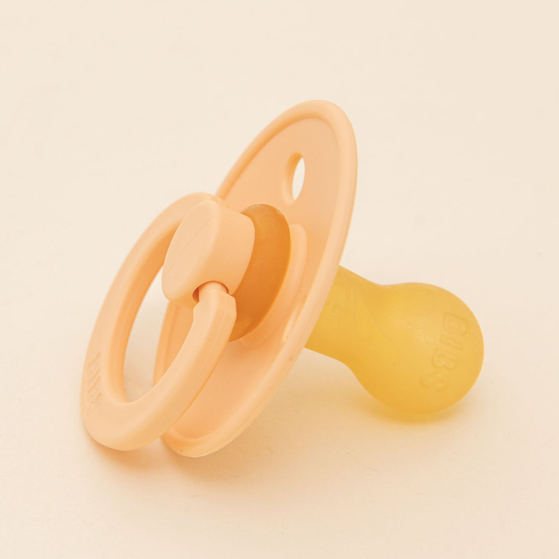 A Bibs Pacifier in Peach Sunset-colored natural rubber baby pacifier on a light beige background. The pacifier features a textured handle and a smooth, rounded shield.