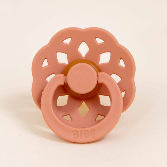 A Bibs Lace Pacifier in Peach with a bohemian inspired design, featuring a circular vented shield and a button-styled handle on a light background.