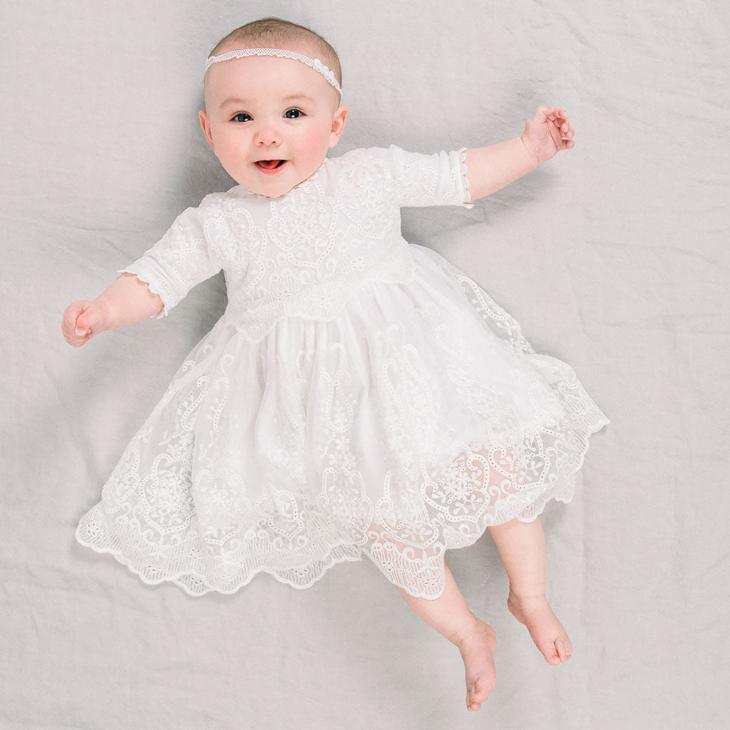 A joyful baby in an Eliza Lace Dress & Headband lies on a light gray background, arms outstretched and smiling.