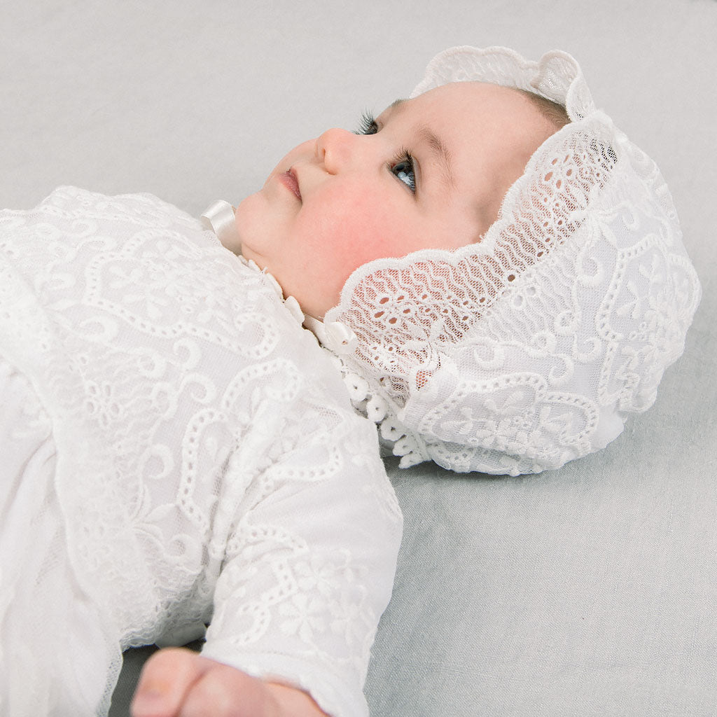 A baby wearing a delicate white lace christening dress and the Eliza Bonnet lies on a soft gray background, looking away thoughtfully.