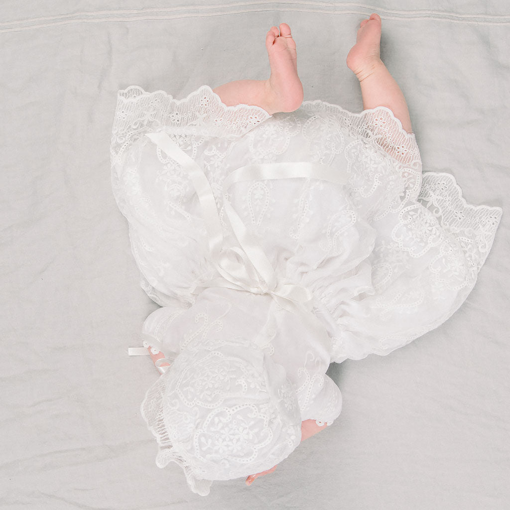 A newborn baby lies on a soft gray fabric, wrapped in an Eliza Lace Dress & Headband with ribbon ties, only the baby’s legs and toes are visible.