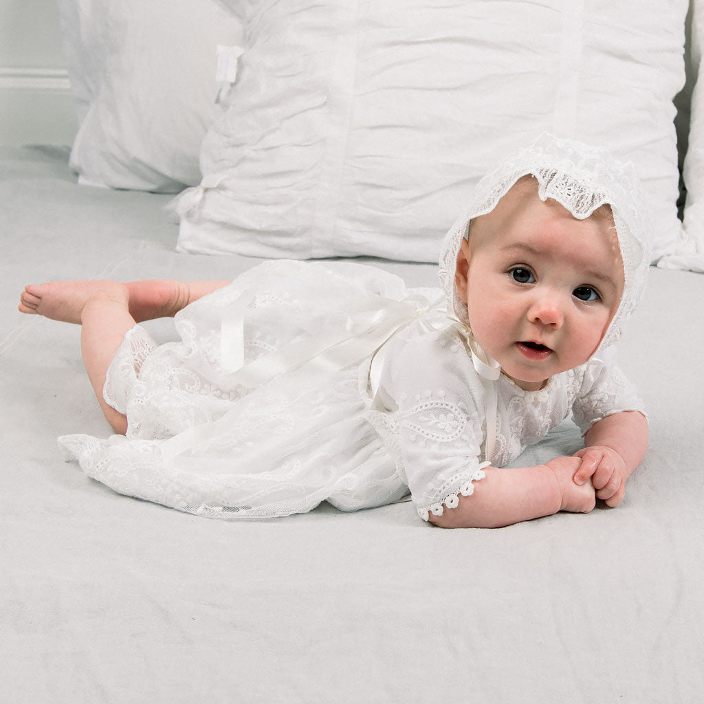 A baby wearing the Eliza Lace Dress & Headband lies on a soft white surface, looking up with a curious expression, surrounded by white pillows, ready for a christening.
