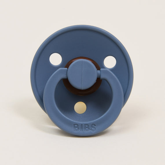 A Bibs Pacifier in Petrol that is blue with a brown handle and the word "bibs" written on it, set against a light beige background.