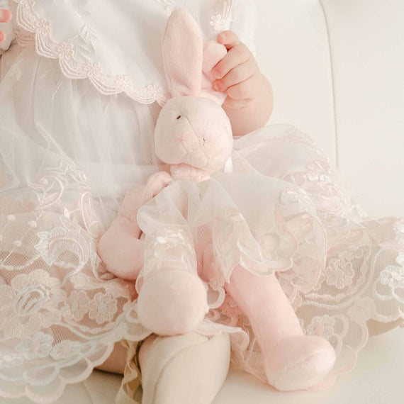 A close-up of a baby holding a Joli Silly Bunny Buddy Pacifier Holder, with focus on the toy and the baby's hands, all against a soft, off-white background.