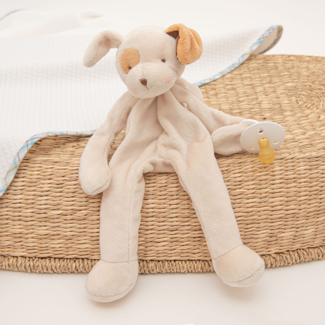 A Mason plush toy puppy, a white and light brown color scheme, sitting on a woven mat next to a baby blanket and holding a pacifier, perfect for a baptism.