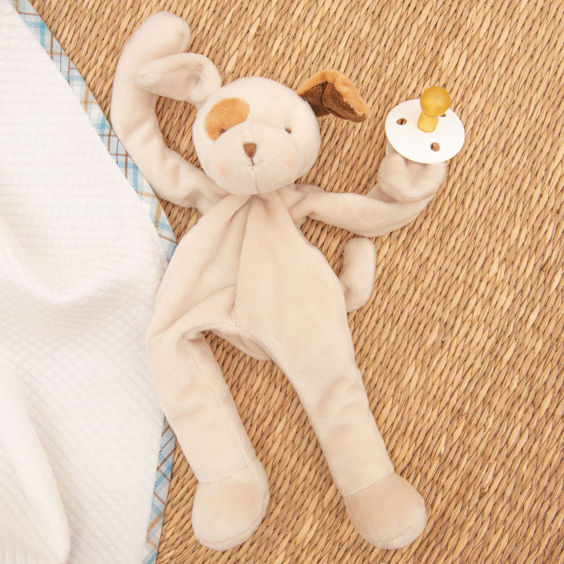 A Mason Puppy Buddy toy and Pacifier on a wicker surface, partially covered by a white baby blanket with a blue patterned trim.