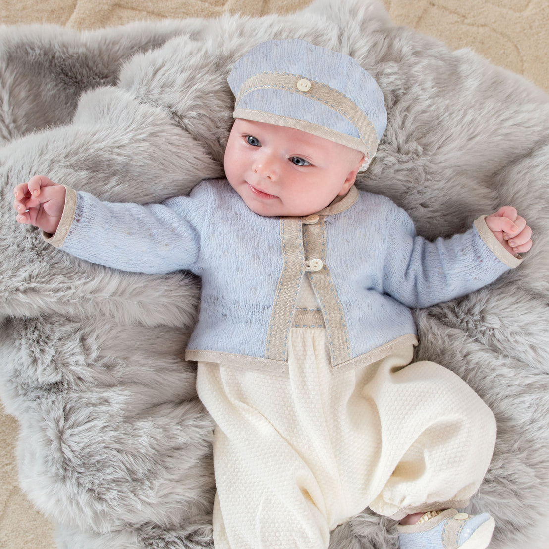 A smiling baby wearing an Austin Sweater and matching cap lies comfortably on a soft grey fur blanket.