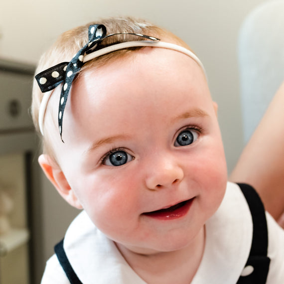 A close-up of a smiling baby with bright blue eyes and a June Bow Headband, wearing a white outfit.