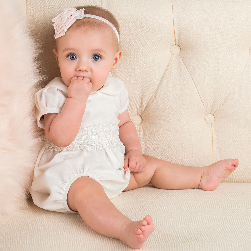 A newborn with blue eyes and light hair, wearing a white outfit for a baptism, sits on a beige sofa looking intrigued, with one hand near her mouth wearing the Emma Linen Rose Headband.