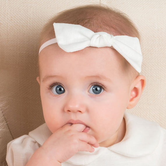 Close-up of a newborn with big blue eyes, wearing an Emma Linen Knot Headband and sucking on their fingers.