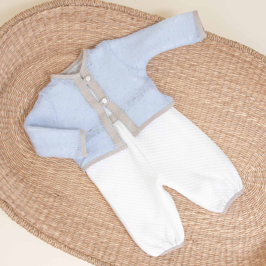 An heirloom baby's coming home two-piece outfit, consisting of an Austin Sweater and white textured pants, displayed on a round rattan mat.