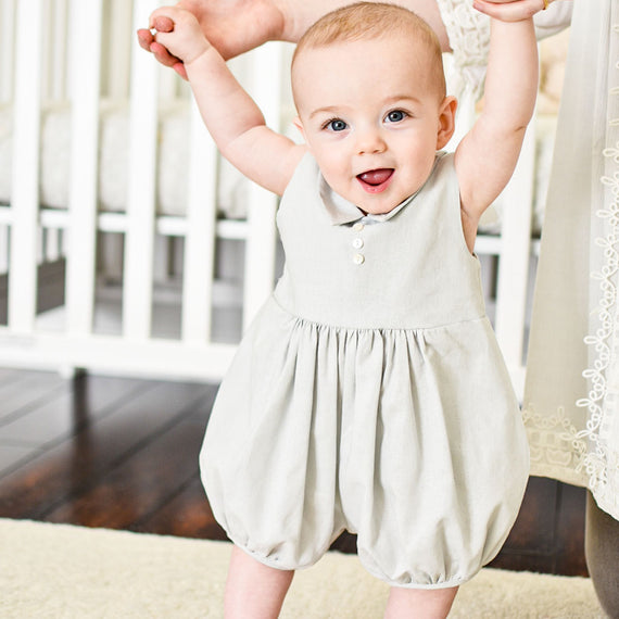 Baby boy held up by his mother. He is wearing the Grayson Linen Romper, a bubble romper made from a soft linen cotton blend in grey and features a pointed collar and button detail on the front.