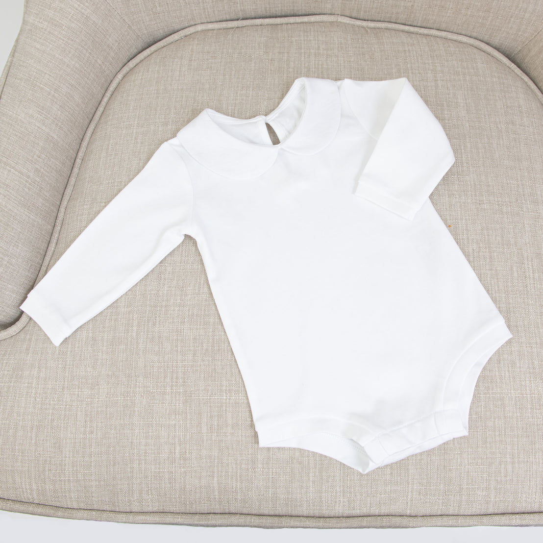 The white onesie, included in the Isla Bubble Skirt Set, lies flat on a textured beige chair.