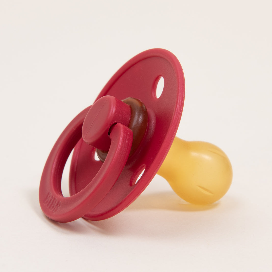 A Bibs Pacifier in Ruby with a natural translucent yellow nipple, set against a plain white background. The handle of the pacifier is ring-shaped and upscale.