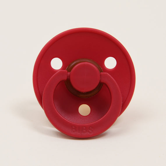 A Bibs Pacifier in Ruby with a button handle, displayed against a plain white background, perfect as an upscale baby gift.