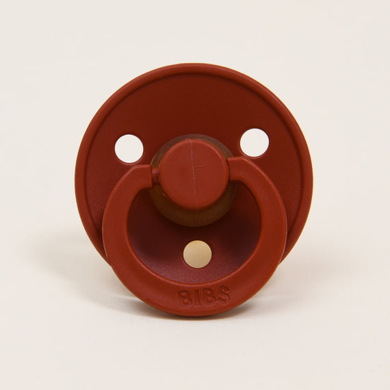 A red Bibs Pacifier in Rust, featuring two air holes and a protruding nipple, set against a plain light background, perfect as an upscale baby gift.