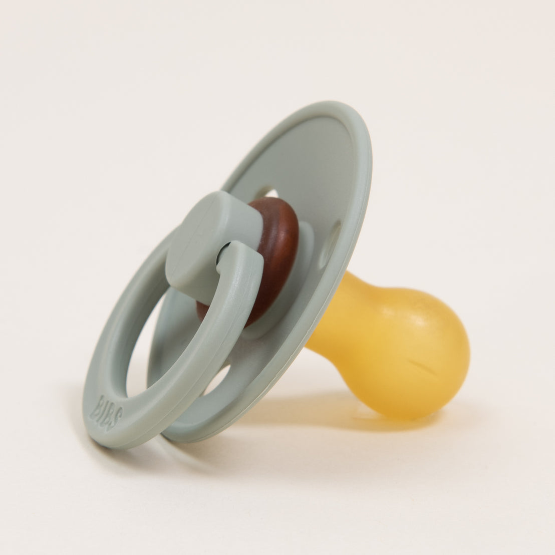 A Bibs pacifier with a pale green handle and yellow nipple, placed on an upscale white background. The pacifier is lying on its side, and the handle is partially loop-shaped.
