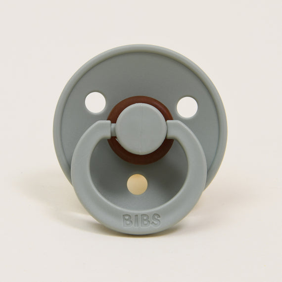 A gray Bibs pacifier with two large air holes and a brown button-style handle, labeled "bibs" on a white background, perfect as an upscale baby gift.