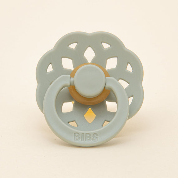 A Bibs Lace pacifier with a sage-colored, flower-shaped silicone shield and a natural rubber button-style center on a beige background.